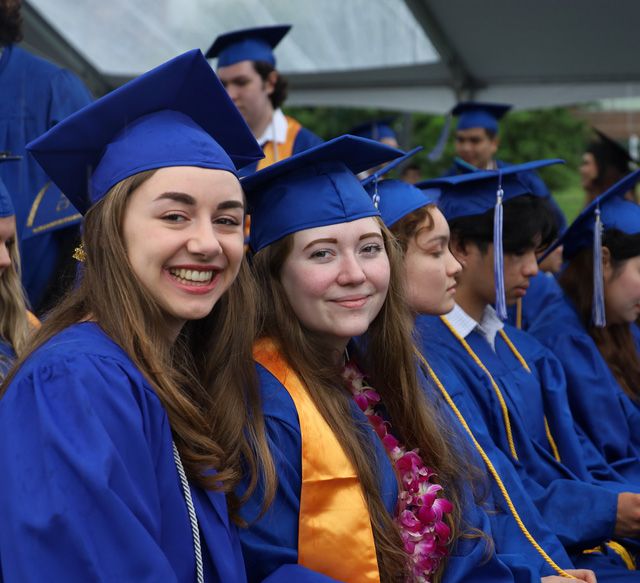 a row of sitting students in graduation regalia with two facing the camera and smiling