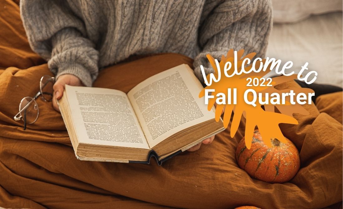 Student reading a book on their bed without their glasses and cozied up in their fall themed bed while wearing a sweater