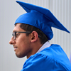 A graduating student wearing blue mortar and gown and glasses looking into distance in front of grey wall