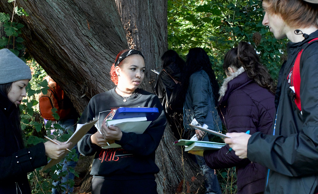 Three people in foreground holding books and discussing notes with four people with backs turned in the wooded background