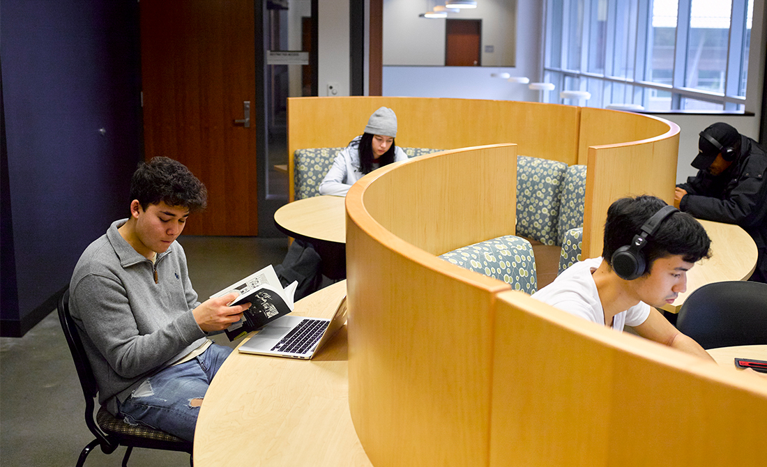 Four students in study cubicles, one reading book with laptop open, two with headphones, and one in a beanie.