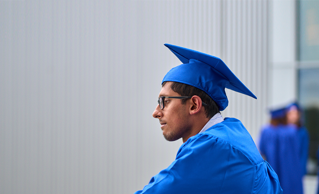 A graduating student wearing blue mortar and gown and glasses looking into distance in front of grey wall