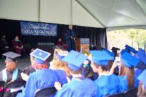 Graduates in blue gowns and caps sitting and watching the college president speak at the ceremony