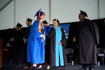Graduate in blue gown elbow bumping with a board of trustee member while collecting her diploma on the graduation stage