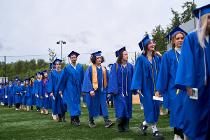 Cascadia graduates in blue gowns and caps walking in a line on the field