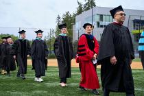 UWB Chancellor in a red gown and others in black gowns walking on the field towards the ceremony