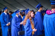 Graduates in blue gowns and caps standing in their seating area under tent